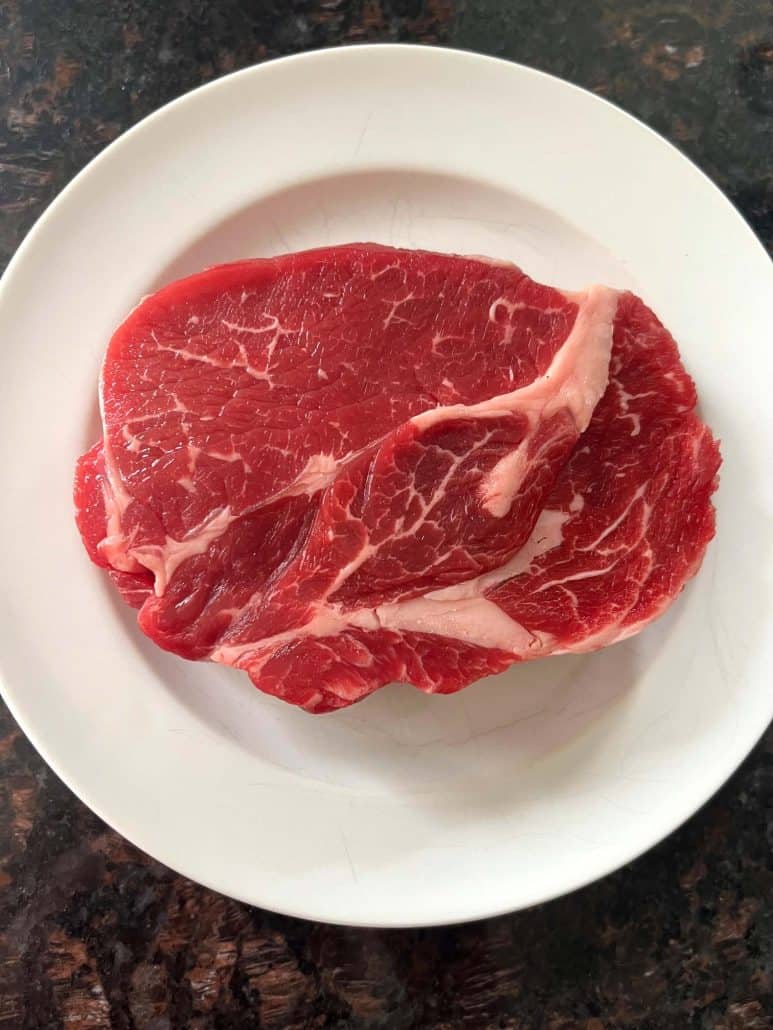 Raw Delmonico steak on a white plate, ready to be seasoned before air frying.