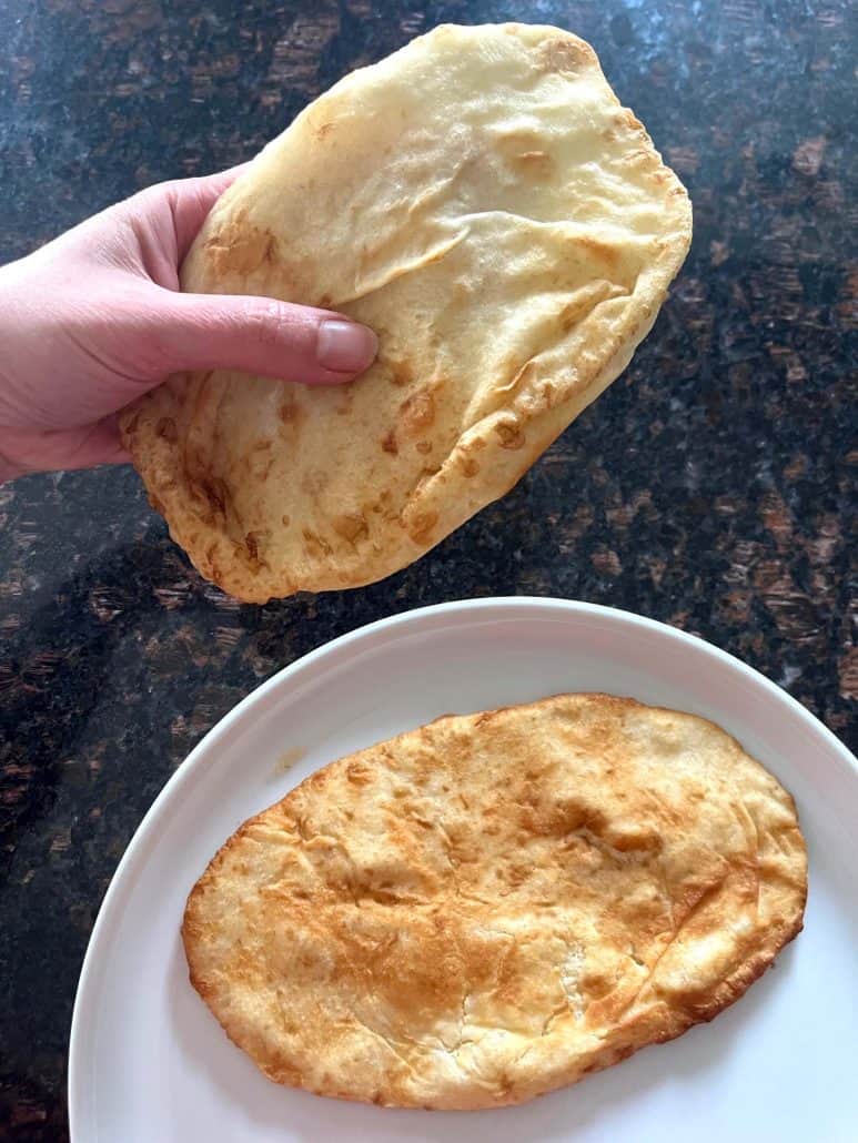 Hand holding a freshly air-fried bhatura above a plate with another piece—showcasing the golden, puffy texture.