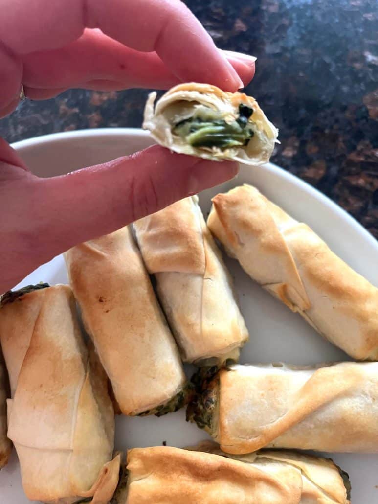Hand holding a halved borek bite to reveal the melted cheese and spinach filling inside, with more golden pastries on a plate below.