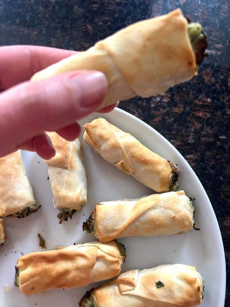 Hand holding a crispy air-fried borek bite over a plate of more borek, showing the flaky texture and spinach filling from the Air Fryer Frozen Borek Bites recipe.