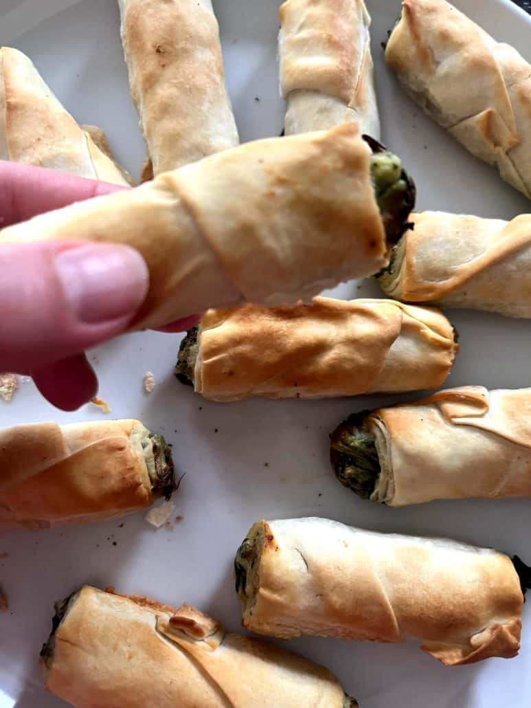 Close-up of a hand holding a golden borek bite above a plate of more pastries, showcasing the flaky phyllo and visible spinach filling from the Air Fryer Frozen Borek Bites recipe.