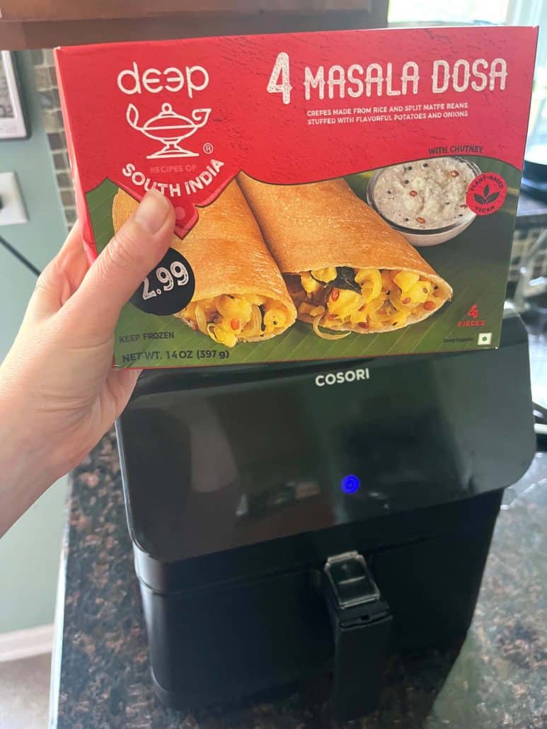 Hand holding a box of frozen masala dosa in front of an air fryer—ready to cook.