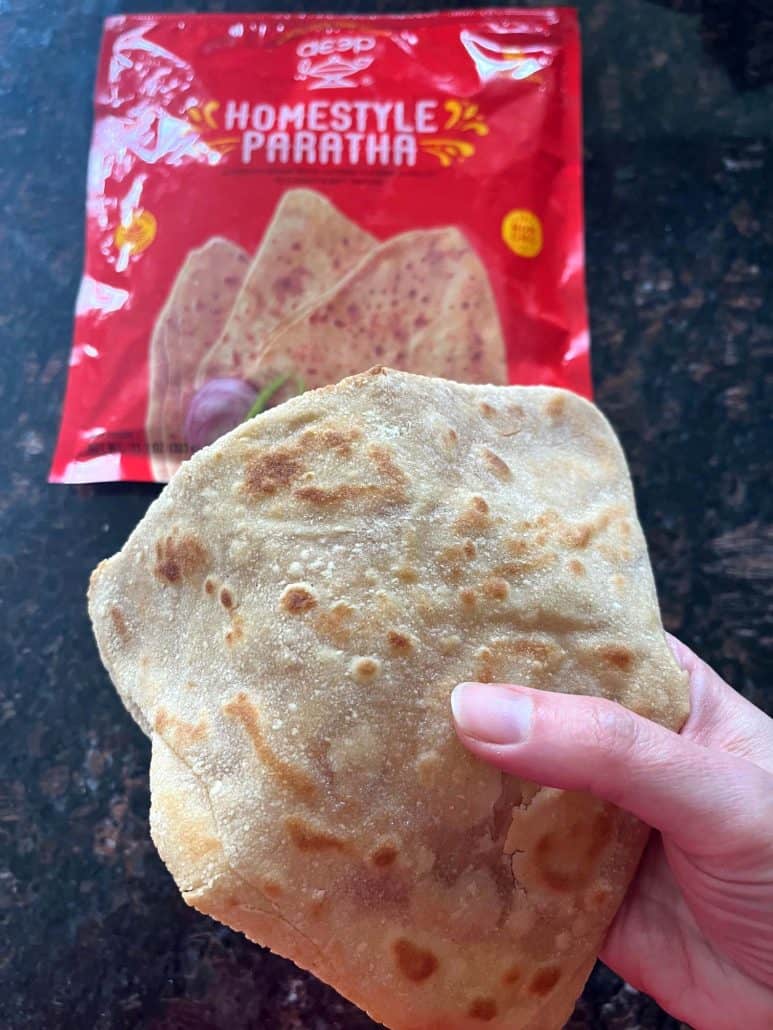 Holding freshly cooked paratha in hand with the frozen paratha packaging in the background—showing the soft, flaky texture after air frying.