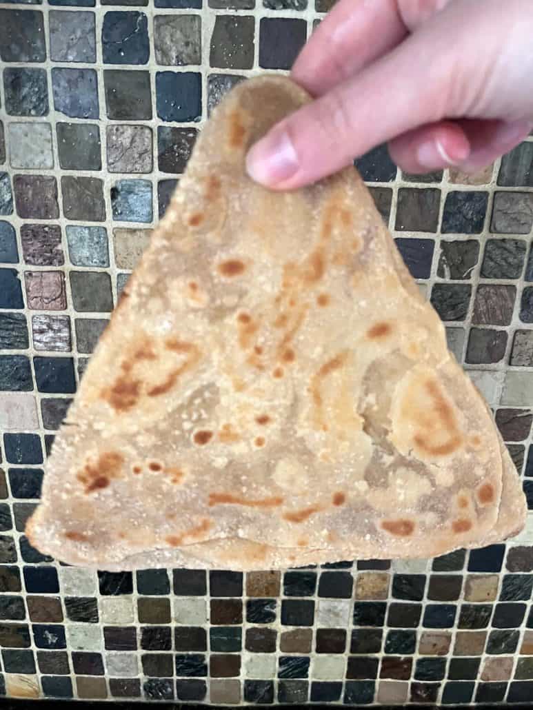 Hand holding a fully cooked paratha in front of a tiled kitchen backsplash—highlighting the golden, flaky texture after air frying.