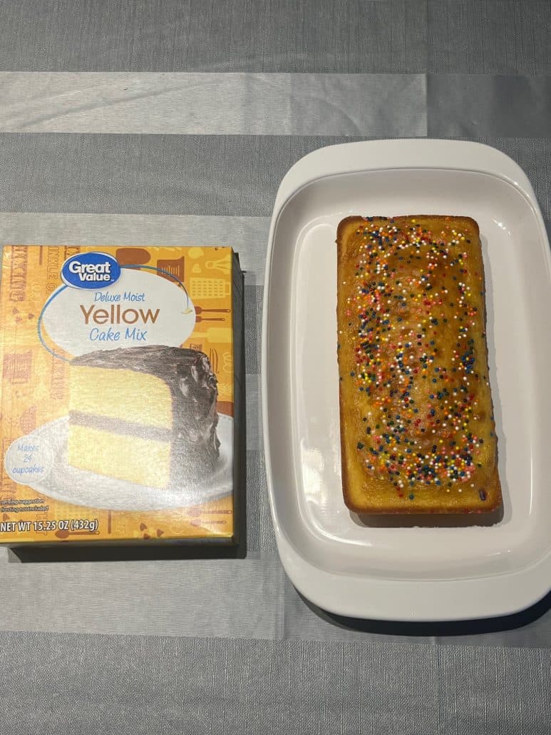 A loaf of cake topped with colorful sprinkles next to a box of cake mix, highlighting the simple ingredients used to make this easy small cake with 1 cup of cake mix.