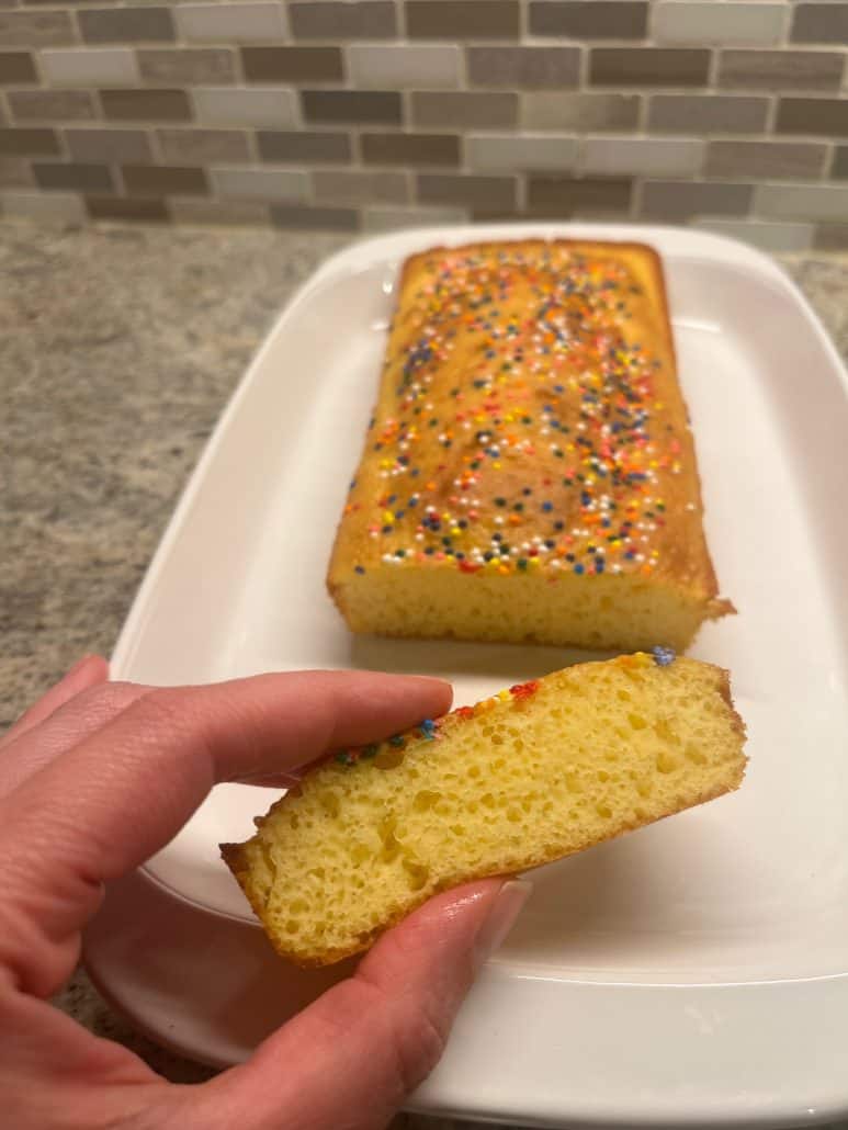 A slice of freshly baked loaf cake being held by hand, revealing a soft interior, with the full cake topped with colorful sprinkles in the background.