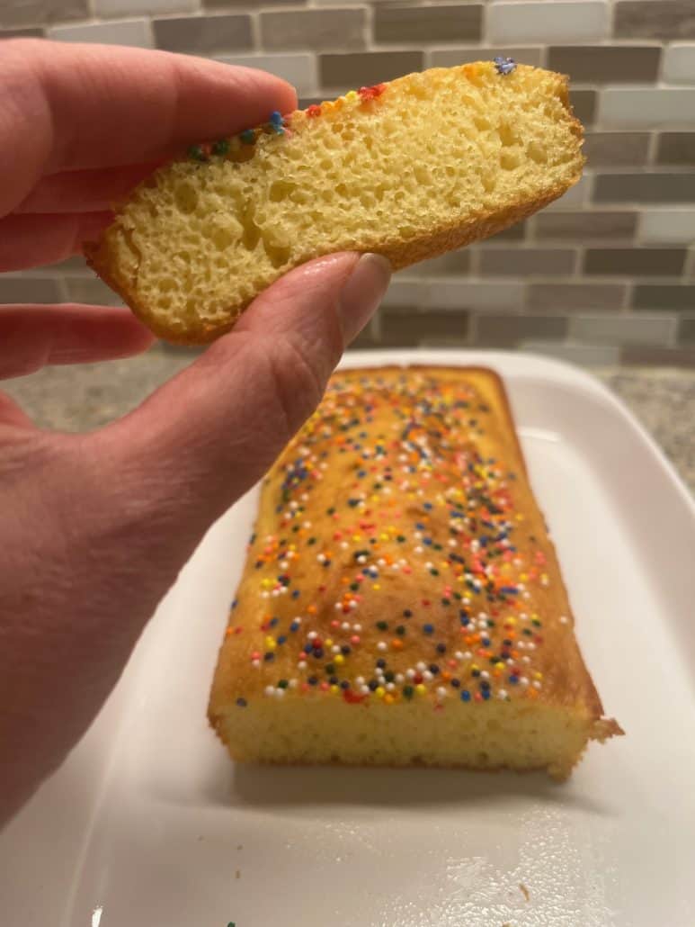 A slice of loaf cake, with the full cake topped with colorful sprinkles displayed in the background.