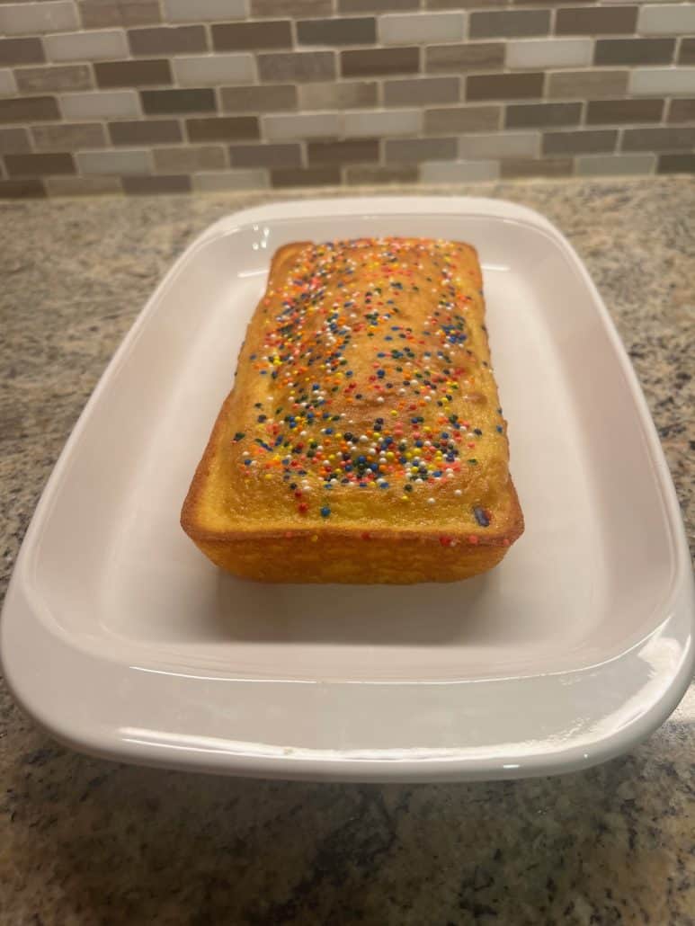 A freshly baked loaf of cake topped with colorful sprinkles, served on a white platter.