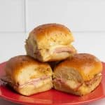 Baked ham and cheese sliders stacked on a red plate, showing their golden tops and melted cheese filling.