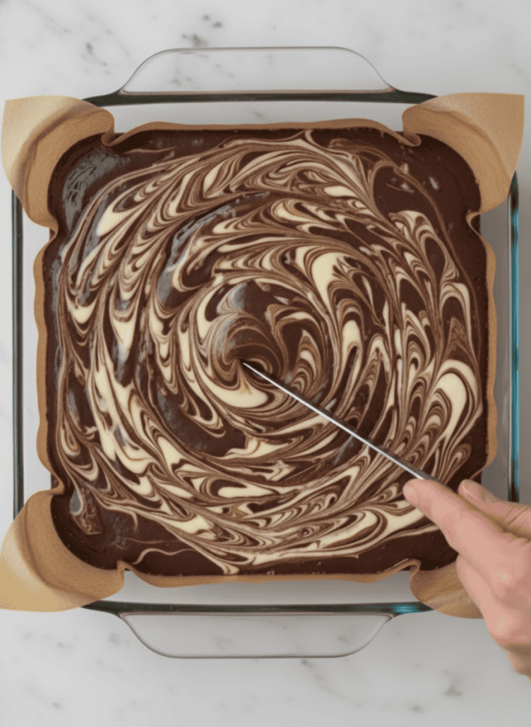 A hand swirling the cheesecake topping into brownie batter in a square baking dish, creating a marbled effect for cheesecake brownies.