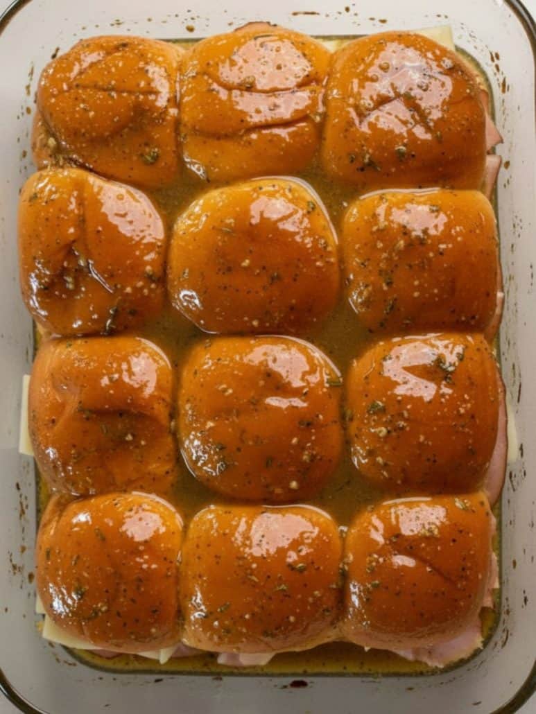 Unbaked ham and cheese sliders in a glass baking dish, topped with golden buns and brushed with a seasoned butter mixture, ready to go in the oven.