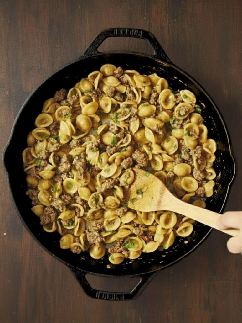 Orecchiette pasta mixed with browned sausage and fresh herbs in a black skillet, stirred with a wooden spoon.