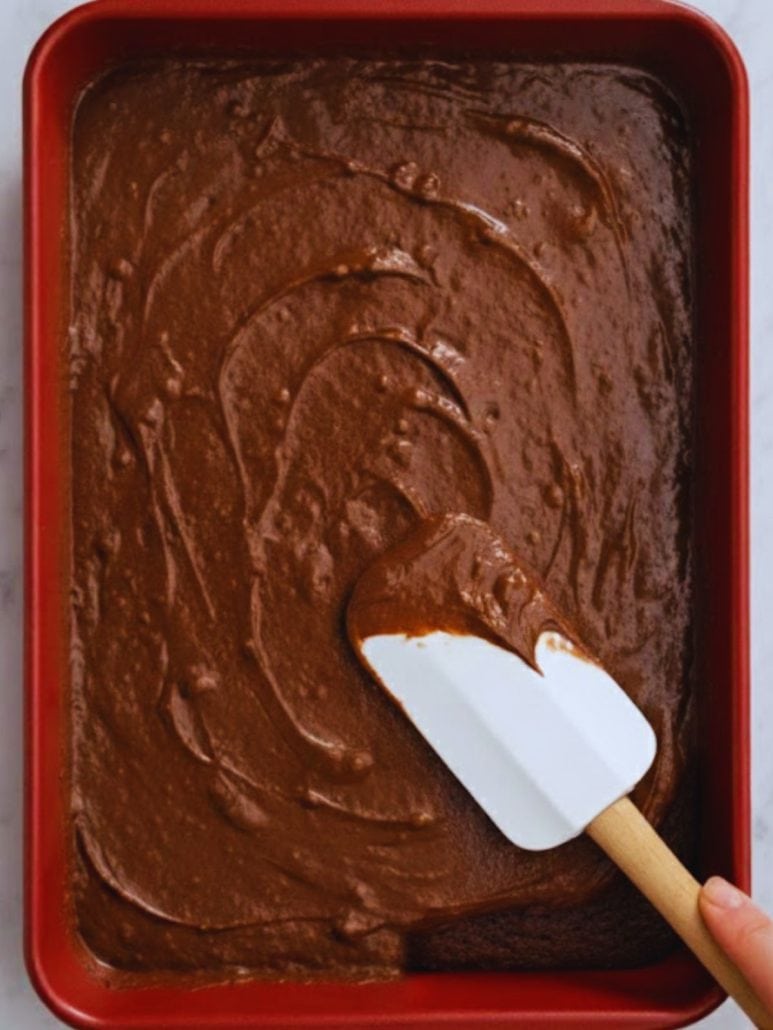 Spreading Texas Sheet Cake batter evenly in a red jelly roll pan using a silicone spatula, just before baking.