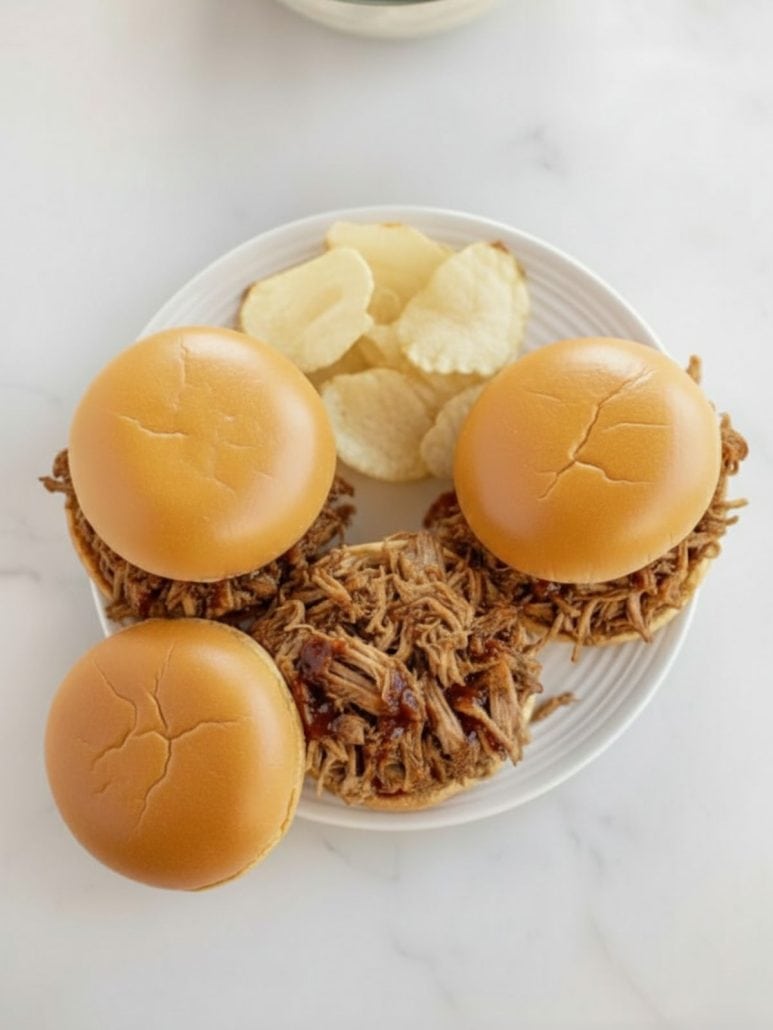 Plate of slow cooker Texas pulled pork sandwiches served with potato chips and extra shredded pork, as featured in the easy pulled pork recipe.