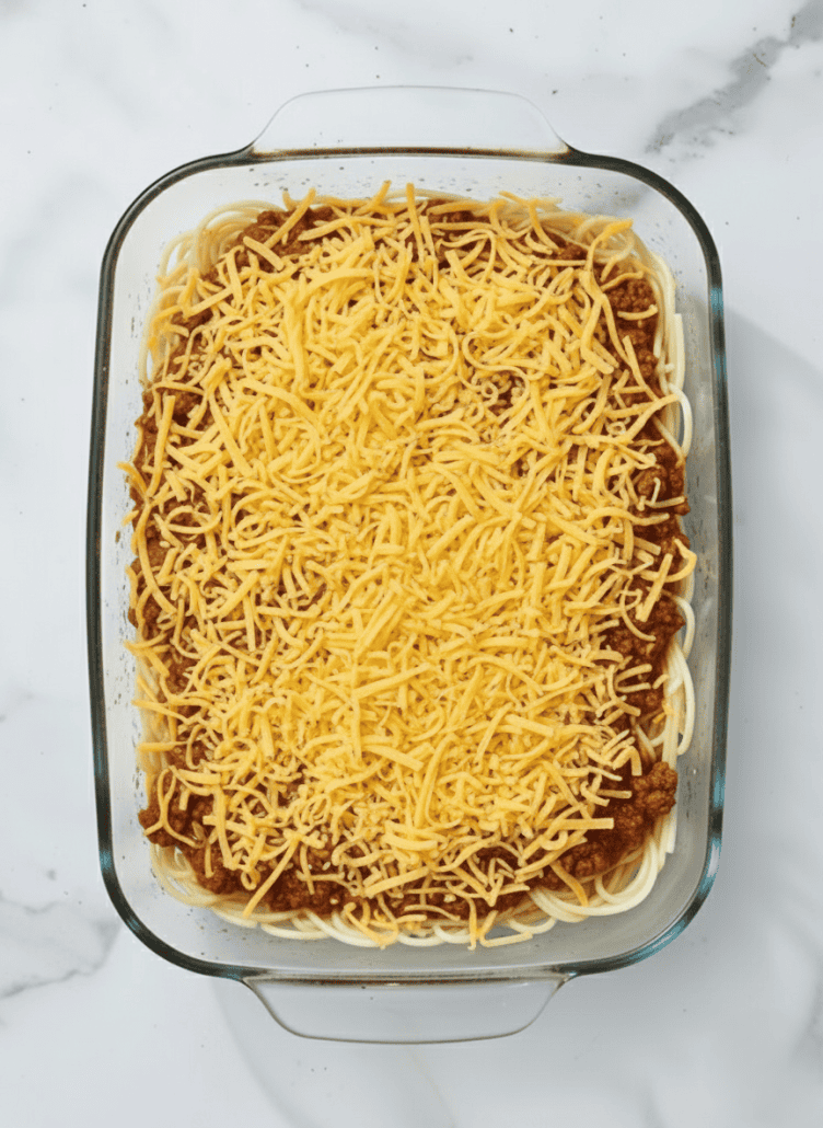 Baked spaghetti with a generous layer of shredded Cheddar cheese on top, ready to be melted and browned in the oven.