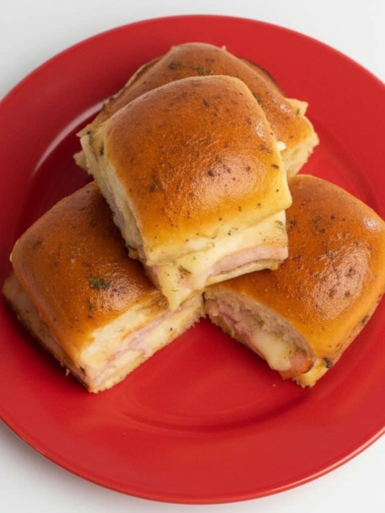A plate of golden brown ham and cheese sliders stacked on a red dish.