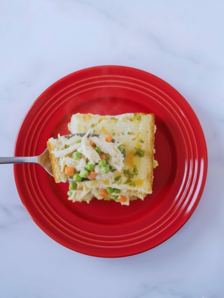 Fork pulling apart a slice of TikTok chicken cobbler on a red plate, revealing creamy shredded chicken, peas, and carrots inside.