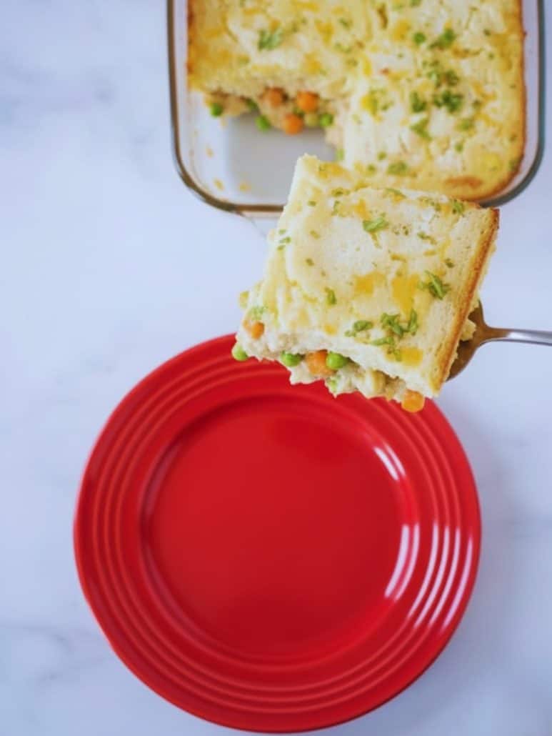 A cheesy slice of TikTok chicken cobbler being served from a baking dish onto a red plate, showing creamy filling with peas and carrots.