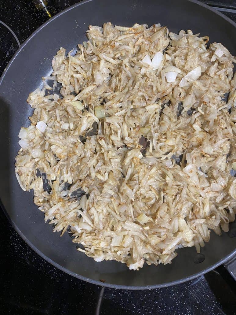 Shredded potatoes and chopped onions sautéing in a skillet. The cooked potato layer for topping the Cheesy Baked Chicken Thighs With Bone In.