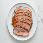 Sliced honey-glazed ham served on a white platter with rosemary, showcasing the caramelized finish.