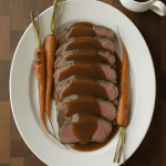 Sliced prime rib roast topped with brown gravy, served with whole roasted carrots on a white platter.