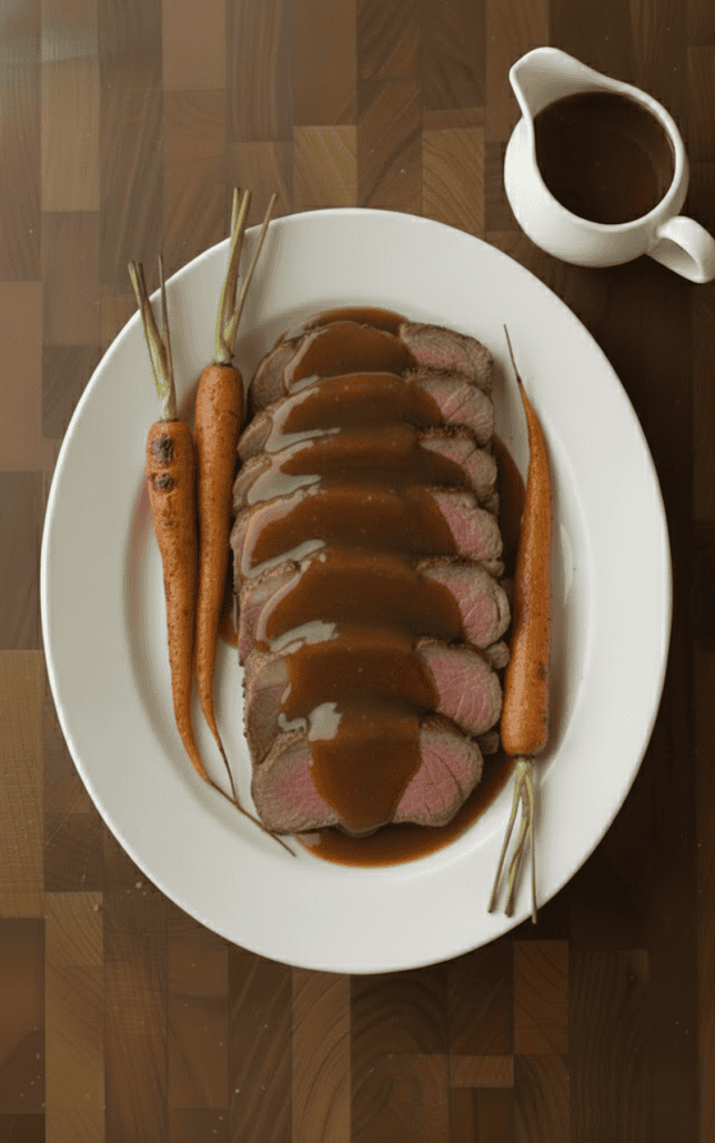 Sliced prime rib roast topped with brown gravy, served with whole roasted carrots on a white platter.