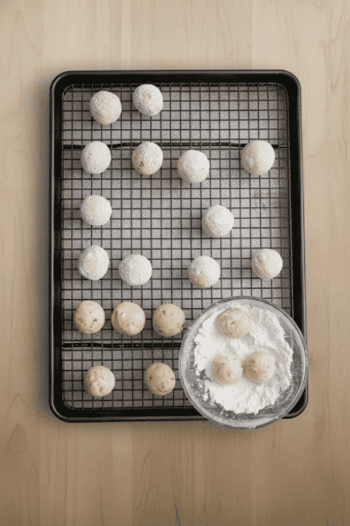 Freshly baked snowball cookies cooling on a wire rack, with a few being rolled in powdered sugar in a shallow bowl—final steps of the holiday cookie recipe.