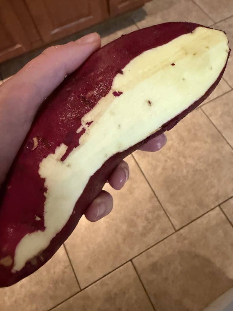 Hand holding a raw white sweet potato with partially peeled red skin, showing its creamy white flesh before roasting.