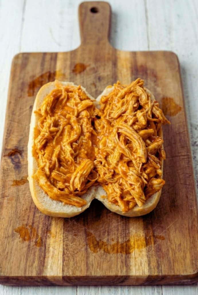 Shredded chicken covered in sauce, placed on a sandwich bun, set on a wooden cutting board, ready to be served.