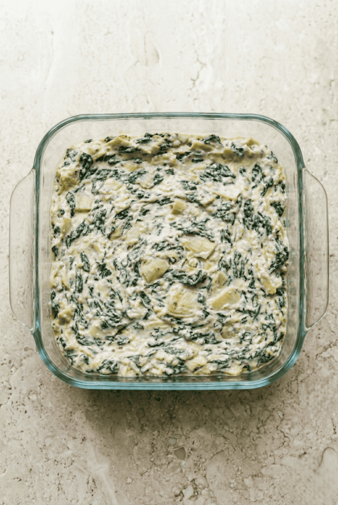 Creamy spinach artichoke dip mixture spread evenly in a glass baking dish. Ready for the next step in the recipe before baking.