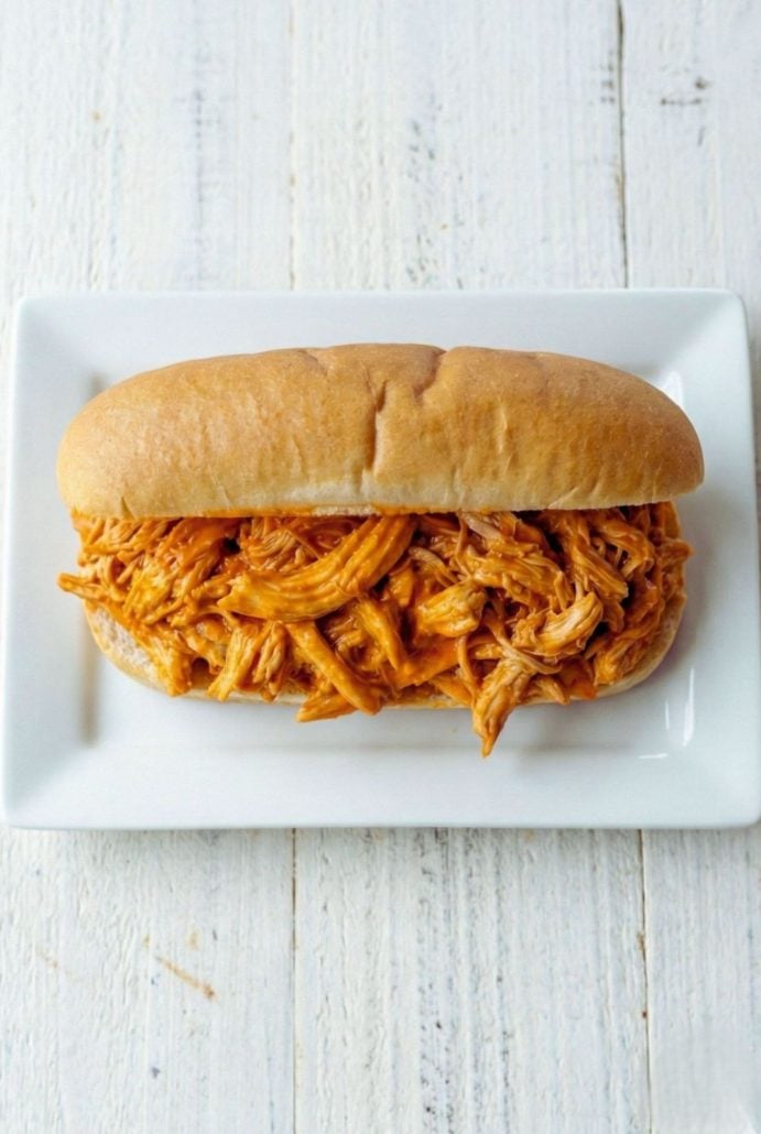 Pulled chicken sandwich on a soft hoagie roll, served on a white plate, showcasing tender shredded chicken in a flavorful sauce.