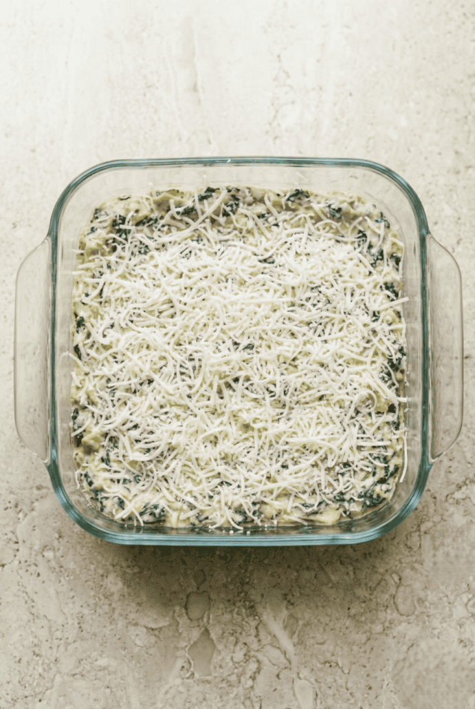 Unbaked spinach artichoke dip in an 8x8 glass baking dish, topped with shredded cheese. Ready to go in the oven.