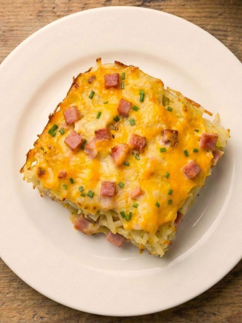 Single serving of ham and hash brown casserole on a white plate, topped with melted cheddar cheese and diced ham.