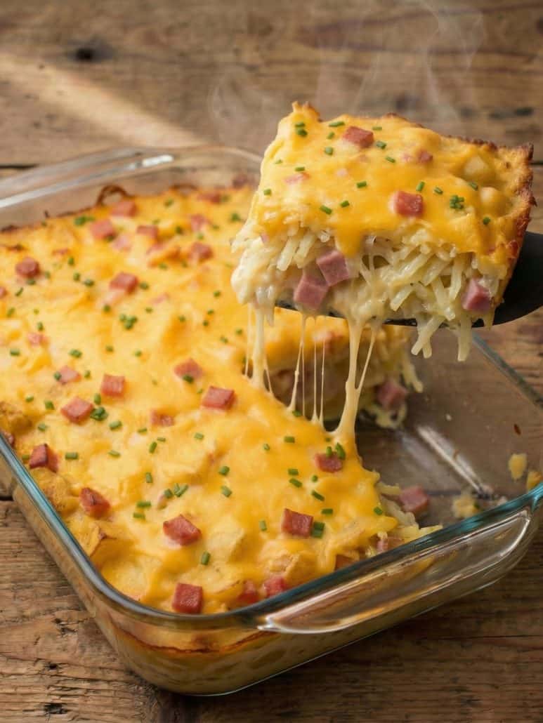 Hot slice of cheesy ham and hash brown casserole being lifted from a baking dish, showing melted cheese and diced ham.