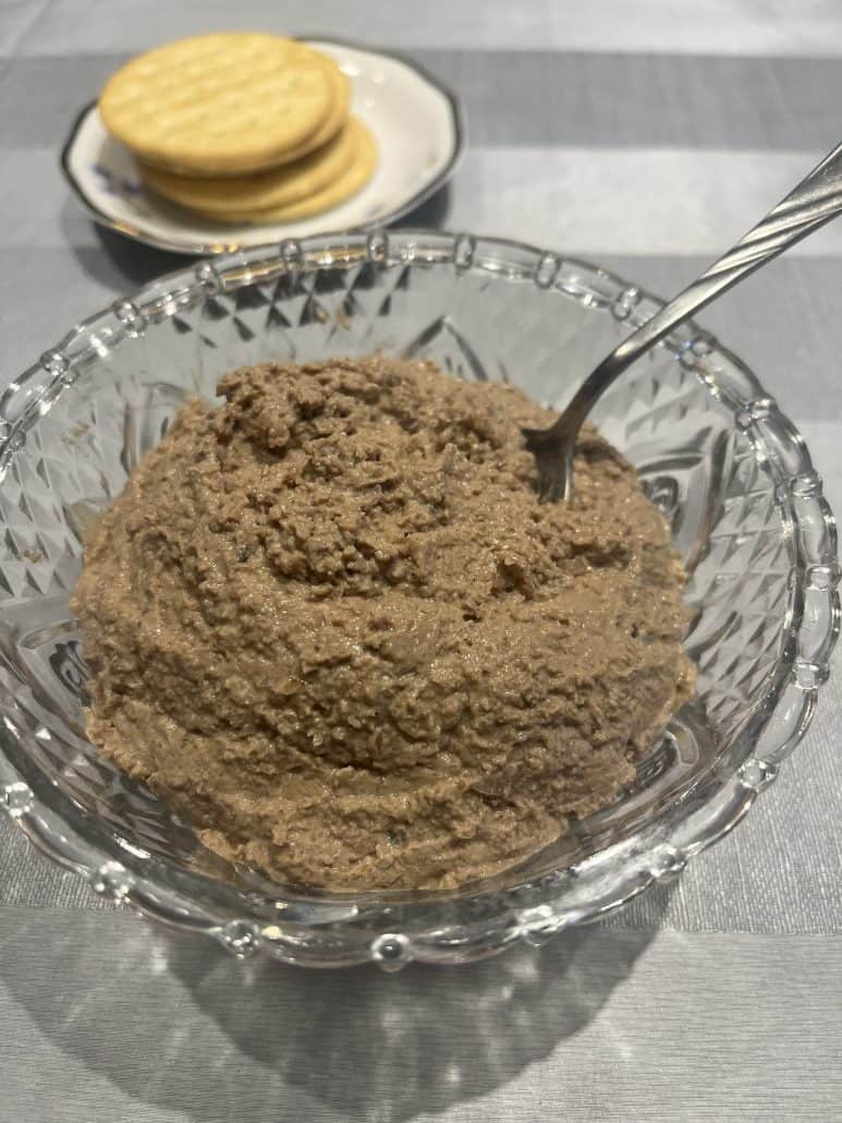 Glass bowl of chopped liver with serving spoon, paired with crackers on a small plate.