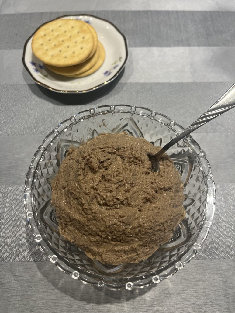 Bowl of homemade chopped liver with a spoon, served alongside a plate of crackers.