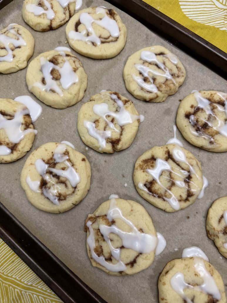 Close-up of freshly baked No Chill Cinnamon Roll Cookies with icing drizzle on a parchment-lined baking sheet.