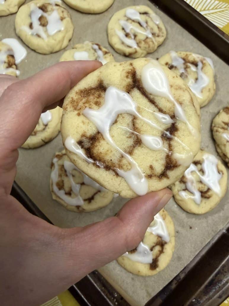 Hand holding a glazed No Chill Cinnamon Roll Cookie above a tray of freshly baked cookies.