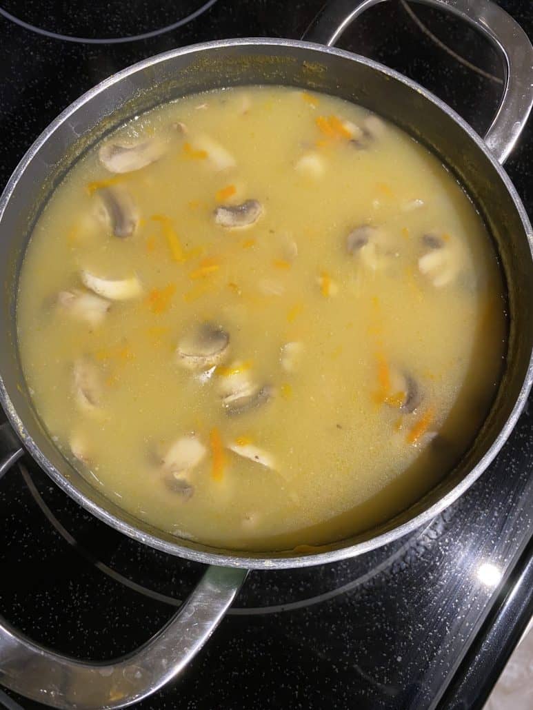 Pot of chunky mushroom soup simmering on the stove, filled with sliced mushrooms and shredded carrots.