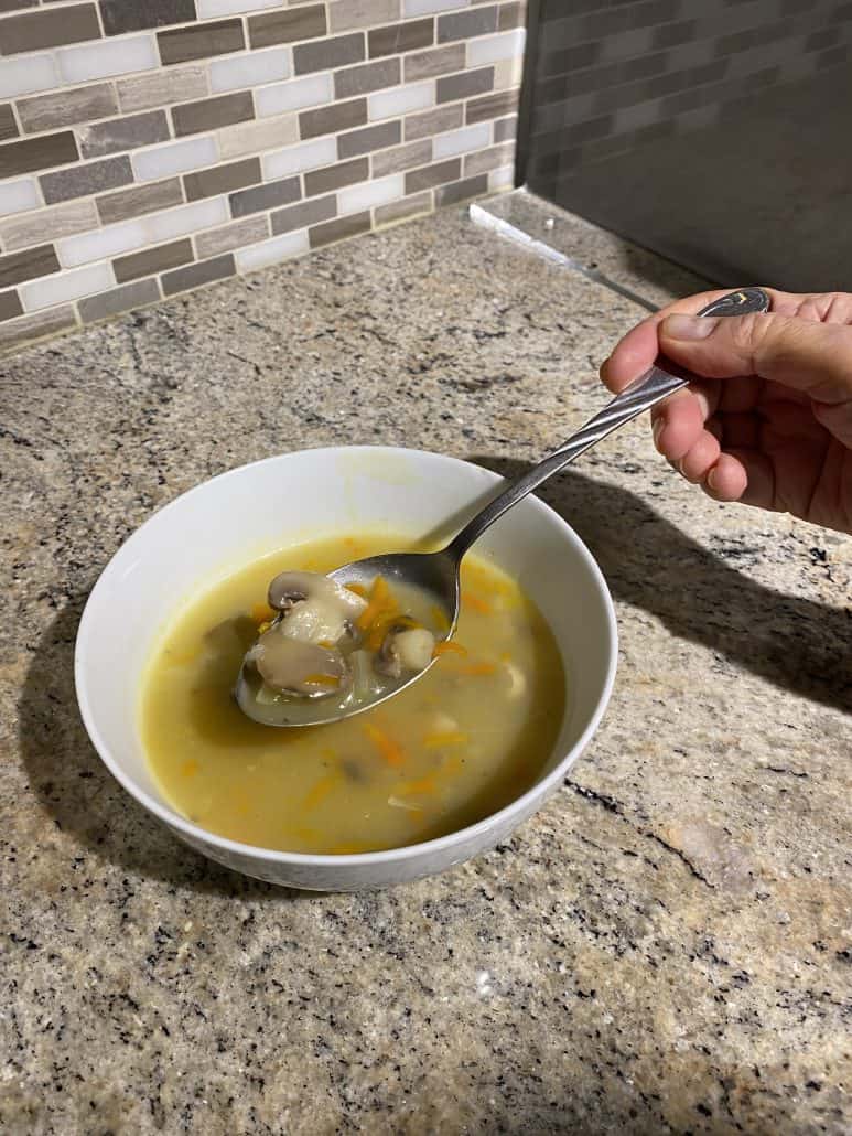Hand holding a spoonful of chunky mushroom soup over a bowl, highlighting the hearty vegetables and creamy texture from the recipe.