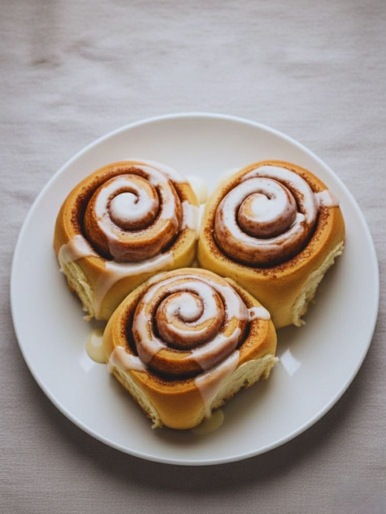 Three homemade overnight cinnamon rolls with glaze on a white plate — a cozy and delicious make-ahead breakfast idea.
