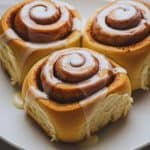 Freshly baked overnight cinnamon rolls topped with vanilla glaze, served warm on a white plate.