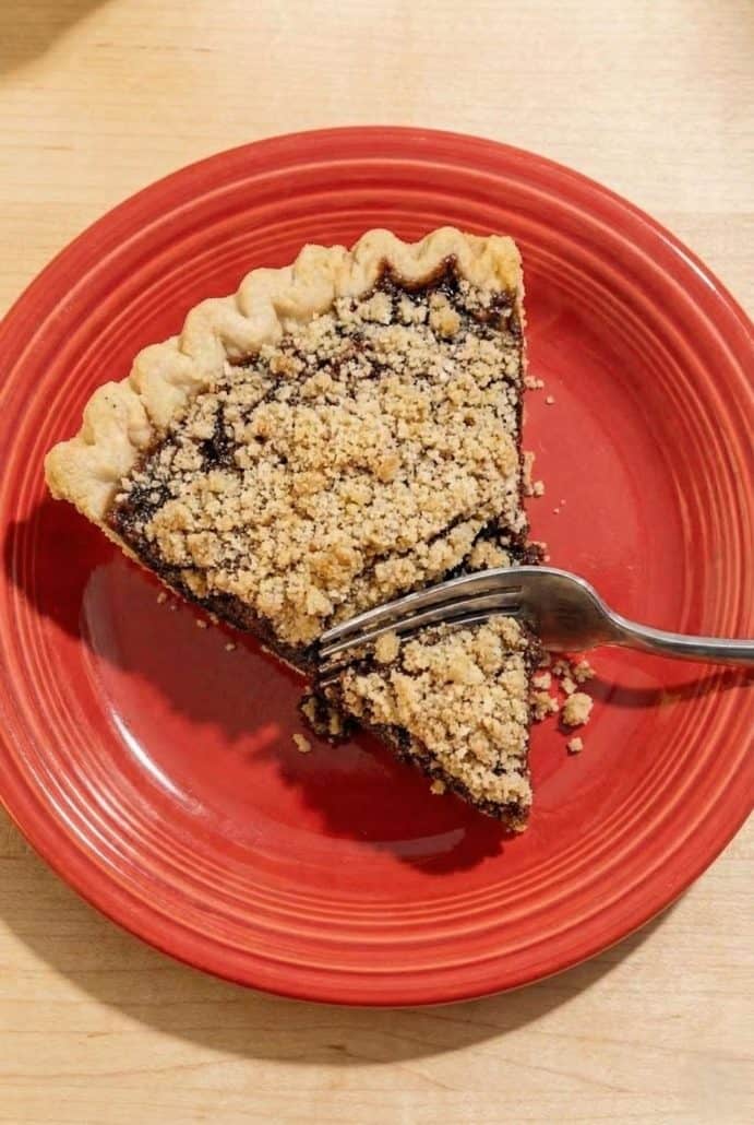 Slice of Shoofly Pie on a red plate with a fork taking a bite, capturing the rich texture of the molasses filling and crumb topping from the featured recipe.