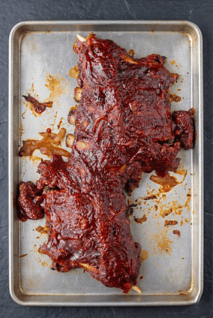Slow cooker baby back ribs coated in BBQ sauce on a baking sheet, ready to be caramelized in the oven.