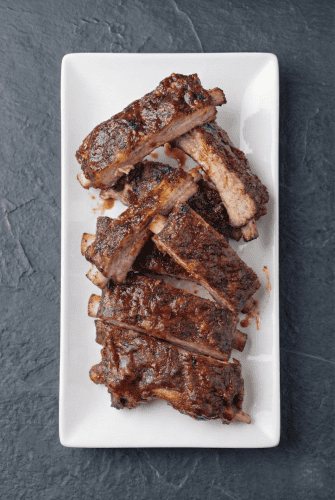 Slow cooker baby back ribs glazed with BBQ sauce on a white platter. Tender, juicy, and caramelized from the oven finish.
