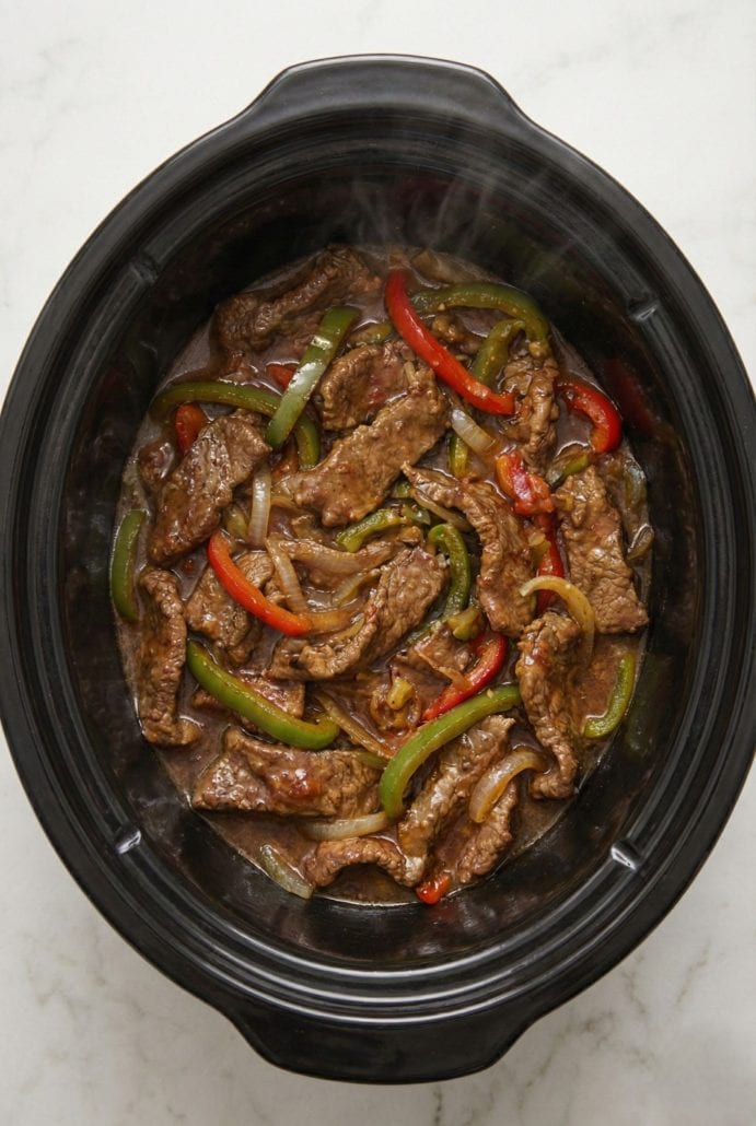Tender pepper steak with bell peppers and onions cooking in a slow cooker. The featured recipe in this easy crockpot dinner post.