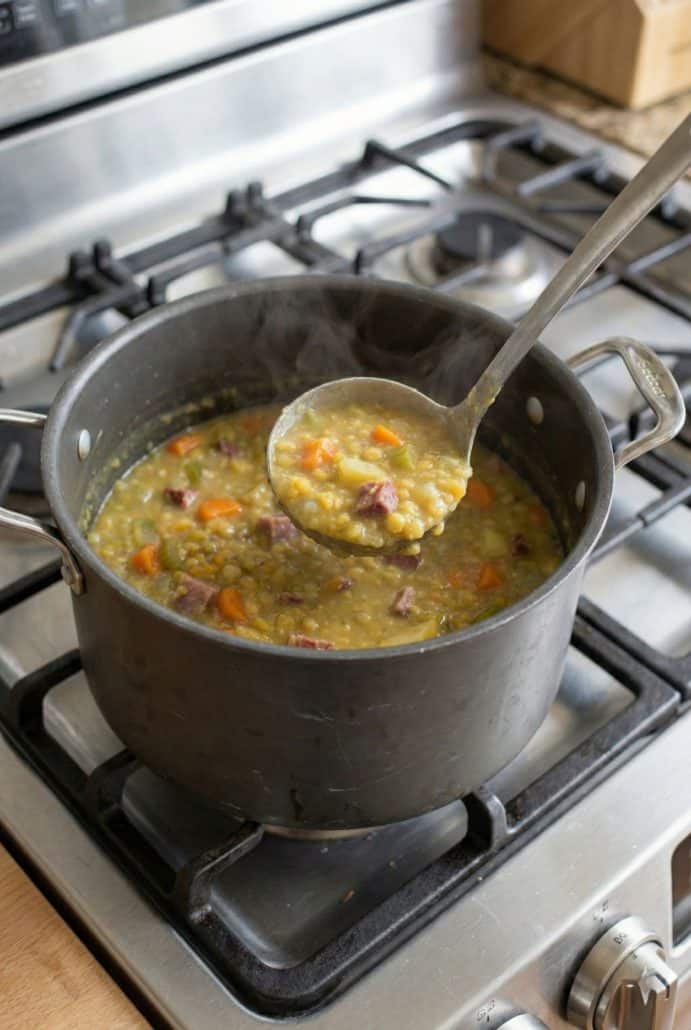 A pot of split pea soup simmering on the stovetop with a ladle full of chunky soup, showing the cooking process for the homemade recipe.