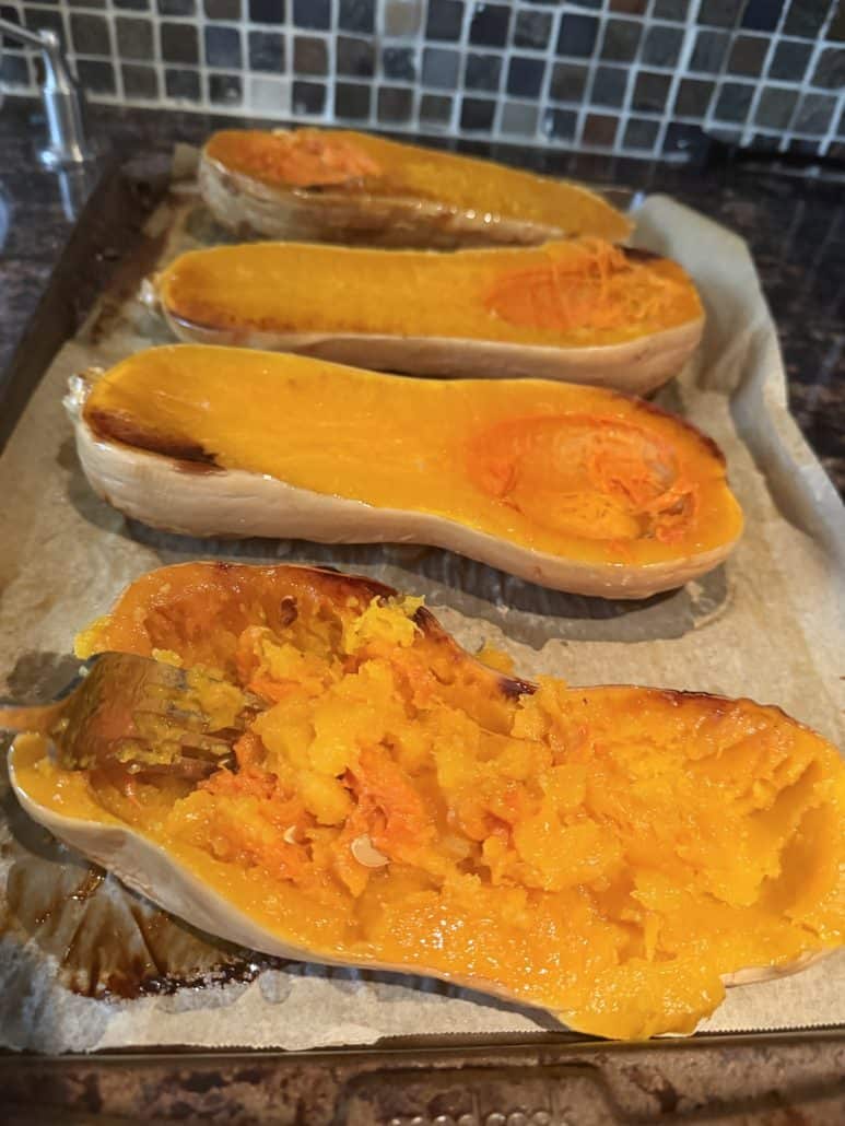 Roasted butternut squash halves on a baking sheet, with one half being scooped out to prepare for vegan stuffing.