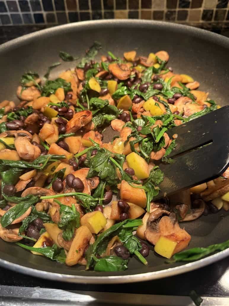 Sautéed mixture of mushrooms, spinach, black beans, and apples in a frying pan for vegan stuffed butternut squash filling.