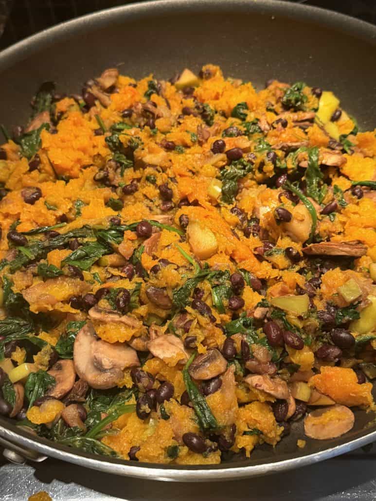 Cooked filling of mashed butternut squash mixed with mushrooms, spinach, black beans, and apples in a skillet, ready to stuff into squash halves.